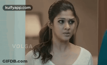 A Woman In A White Shirt Is Standing In Front Of A Glass Door Looking At The Camera . GIF