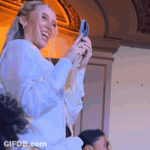 A Woman In A White Shirt Is Taking A Picture With A Cell Phone GIF