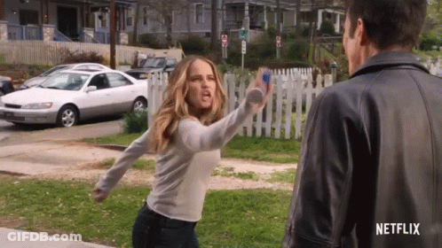 A Woman In A White Shirt Is Throwing A Bottle At A Man In A Leather Jacket . GIF