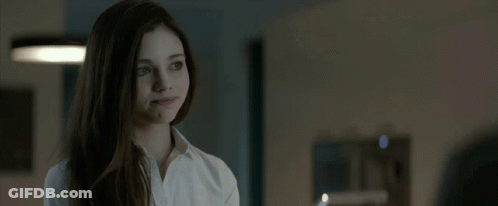 A Woman In A White Shirt Looks At The Camera In A Dark Room GIF