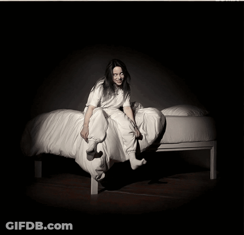 A Woman In A White Shirt Sits On A Bed GIF