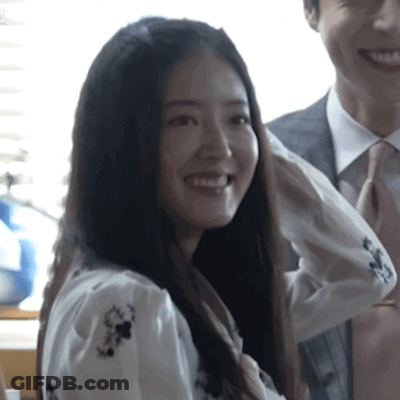 A Woman In A White Shirt With Flowers On It Smiles Next To A Man In A Suit And Tie GIF