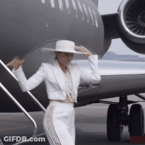 A Woman In A White Suit And Hat Is Standing In Front Of A Plane GIF