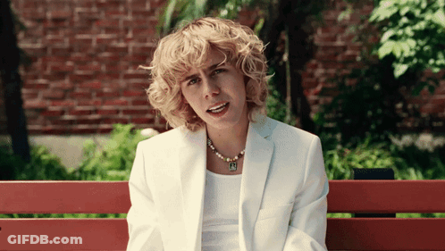 A Woman In A White Suit Is Sitting On A Bench With A Brick Wall In The Background GIF