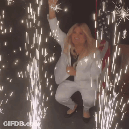 A Woman In A White Suit Stands In Front Of A Firework Display GIF
