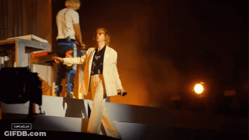 A Woman In A White Suit Stands On A Stage In Front Of A Sign That Says Gascella GIF