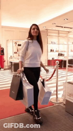 A Woman In A White Sweater And Black Pants Is Carrying Shopping Bags In A Store GIF