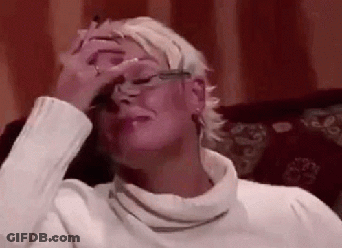 A Woman In A White Sweater And Glasses Is Sitting On A Couch Covering Her Face With Her Hand . GIF