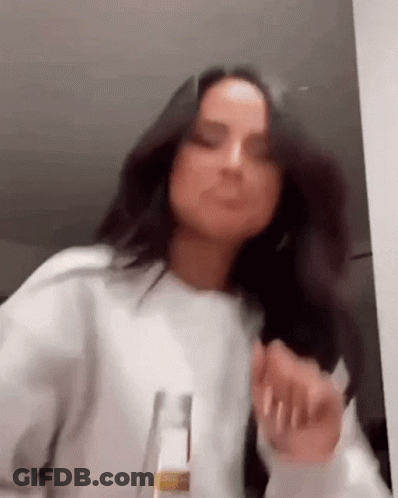 A Woman In A White Sweater Is Dancing In A Room With A Bottle In Her Hand . GIF