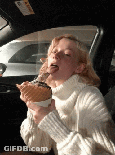 A Woman In A White Sweater Is Eating An Ice Cream Cone In A Car GIF