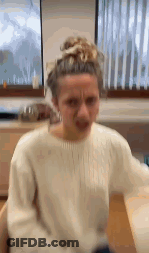 A Woman In A White Sweater Is Making A Funny Face In Front Of A Window . GIF