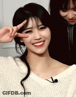 A Woman In A White Sweater Is Smiling And Giving A Peace Sign GIF