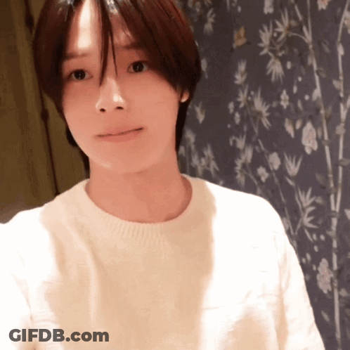 A Woman In A White Sweater Stands In Front Of A Wall With Flowers On It GIF