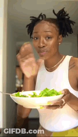 A Woman In A White Tank Top Is Holding A Bowl Of Salad With A Fork . GIF