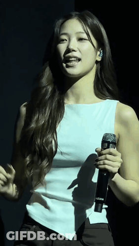 A Woman In A White Tank Top Is Singing Into A Microphone GIF