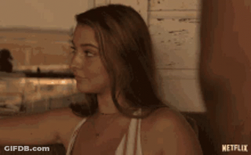 A Woman In A White Tank Top Is Sitting On A Couch And Looking At Something . GIF