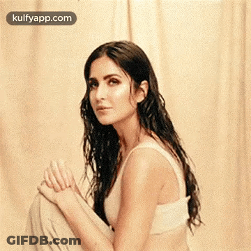 A Woman In A White Tank Top Is Sitting On The Floor With Her Legs Crossed And Looking At The Camera . GIF