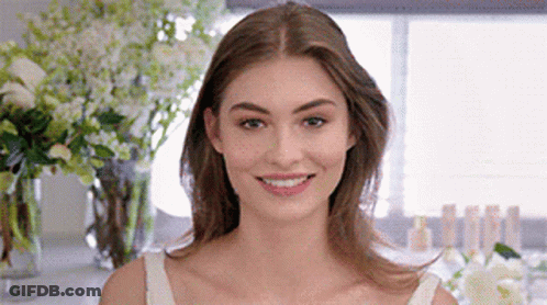 A Woman In A White Tank Top Is Smiling In Front Of Flowers . GIF