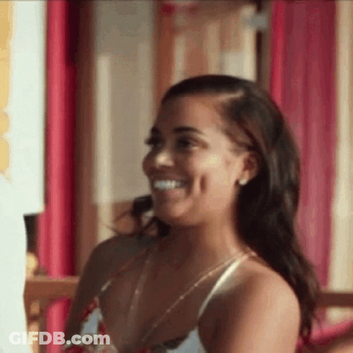 A Woman In A White Tank Top Is Smiling While Sitting In Front Of A Pink Wall . GIF