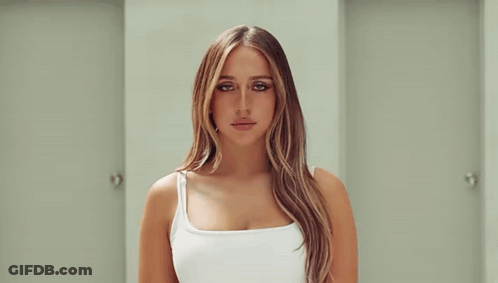 A Woman In A White Tank Top Is Standing In A Hallway Between Two Doors . GIF