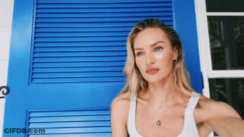 A Woman In A White Tank Top Is Standing In Front Of A Blue Door GIF