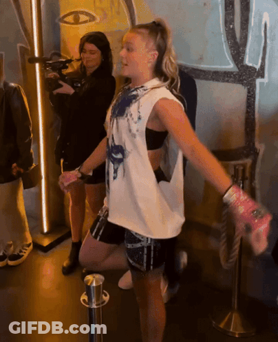 A Woman In A White Tank Top Is Standing In Front Of A Camera GIF