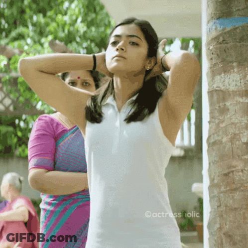 A Woman In A White Tank Top Is Standing Next To A Palm Tree . GIF