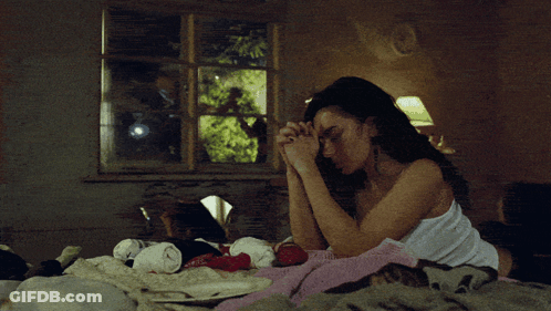 A Woman In A White Tank Top Sits On A Bed With Her Hands Folded GIF