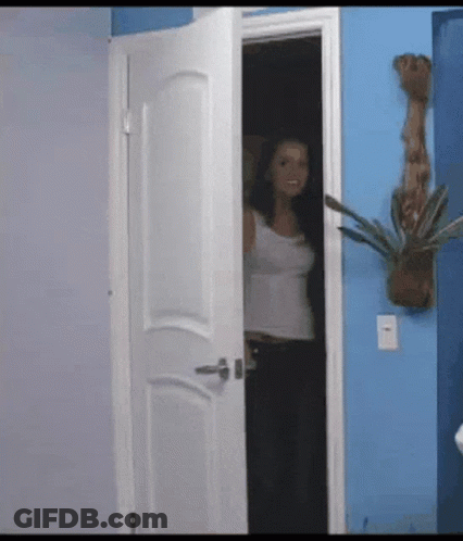 A Woman In A White Tank Top Stands In A Doorway In Front Of A Blue Wall GIF