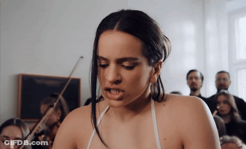 A Woman In A White Tank Top Stands In Front Of A Group Of People Playing Violins GIF