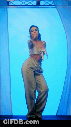 A Woman In A White Top And Grey Pants Is Dancing In Front Of A Blue Curtain GIF