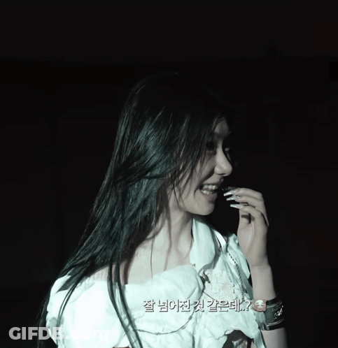 A Woman In A White Top Is Smiling In A Dark Room With Korean Writing On The Bottom GIF