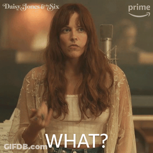 A Woman In A White Top Says " What " While Standing In Front Of A Microphone GIF
