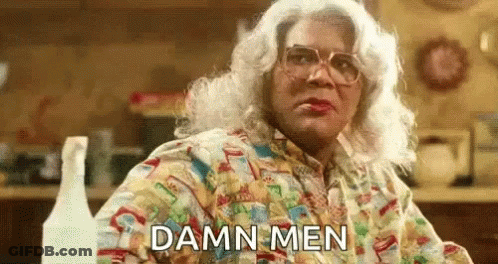 A Woman In A Wig And Glasses Is Sitting At A Table In A Kitchen And Says `` Damn Men '' . GIF