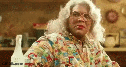 A Woman In A Wig And Glasses Is Sitting At A Table In A Kitchen With A Spray Bottle . GIF