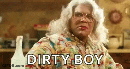 A Woman In A Wig And Glasses Is Sitting At A Table With The Words `` Dirty Boy '' Written On The Screen . GIF