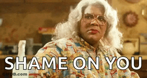 A Woman In A Wig And Glasses Is Sitting At A Table With The Words `` Shame On You '' Written On It . GIF