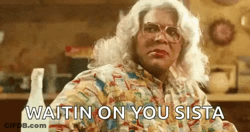 A Woman In A Wig And Glasses Is Sitting At A Table With The Words `` Waitin On You Sista '' . GIF