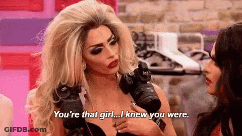 A Woman In A Wig Is Holding A Camera And Talking To Another Woman . GIF