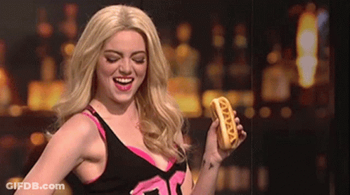 A Woman In A Wig Is Holding A Hot Dog That Says ' Vava ' On It GIF