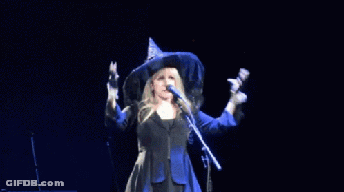 A Woman In A Witch Hat Sings Into A Microphone GIF