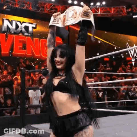 A Woman In A Wrestling Ring Holding A Championship Belt In Front Of A Sign That Says Nxt Venge GIF