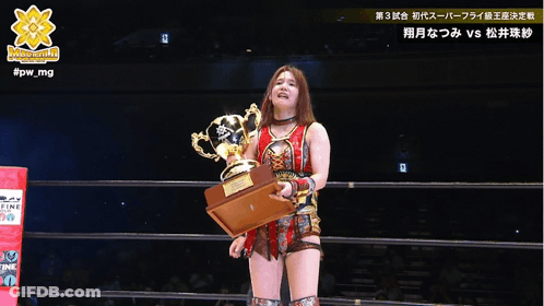 A Woman In A Wrestling Ring Holds A Trophy In Front Of A Referee With #Pw_mg Written On The Bottom GIF