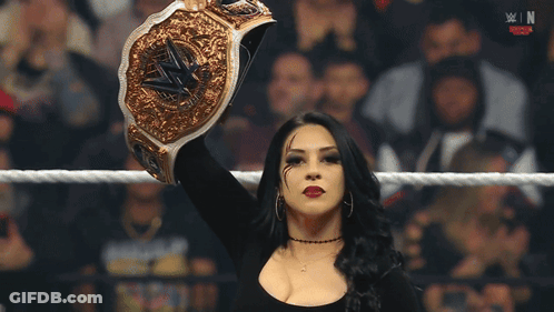 A Woman In A Wrestling Ring Holds Up A Gold Wrestling Championship Belt GIF