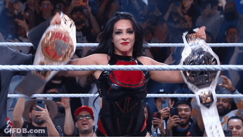 A Woman In A Wrestling Ring Holds Up Two Belts In Front Of A Crowd GIF
