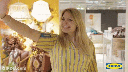 A Woman In A Yellow And Blue Striped Shirt Is Waving In Front Of A Sign That Says Ikea GIF