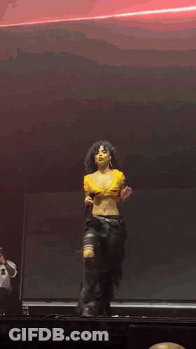 A Woman In A Yellow Crop Top And Black Pants Is Dancing On A Stage GIF