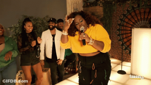 A Woman In A Yellow Crop Top Singing Into A Microphone In Front Of People GIF