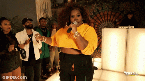 A Woman In A Yellow Crop Top Sings Into A Microphone In Front Of A Group Of People GIF