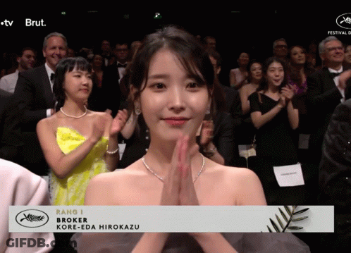 A Woman In A Yellow Dress Applauds In Front Of A Banner That Says Rang J Broker Kore-Eda Hirokazu GIF